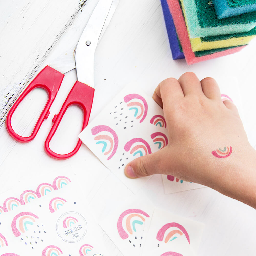 Kind greift zu den Klebetattoos Kleiner Regenbogen die auf einem weißen Tisch liegen